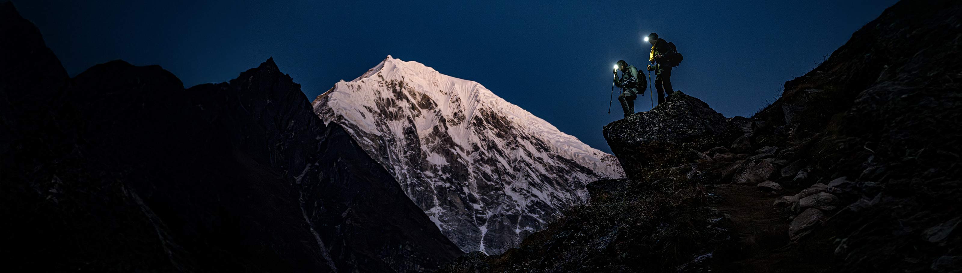 Two hikers wearing headlamps standing on a rocky ledge of a mountain at night